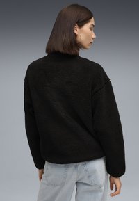 Pullover in pile nero con superficie texturizzata e vestibilità ampia, caratterizzato da un collo alto e spalle scese. Abbinato a jeans azzurri chiari.