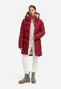 Czerwona puchowa kurtka z kapturem, z teksturowanej tkaniny, wyszywana, z brązowym futrzanym wykończeniem. Zestawiona z białymi spodniami i taupe kozakami.