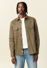 Camicia verde oliva con bottoni, dotata di due tasche sul petto, con colletto a punta, indossata sopra una maglietta bianca a girocollo e jeans azzurri chiari.