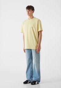 Geltona medvilninė marškinė su apvaliu kaklu ir trumpomis rankovėmis, derinama su šviesiai mėlynomis plačiomis kelnėmis ir juodais sportbačiais. Paprastas dizainas, be raštų.