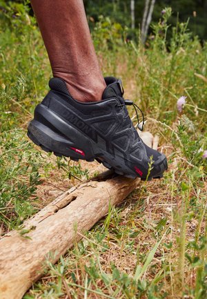 Zwarte trailrunning schoen met een gestructureerde bovenkant en een robuuste zool, zittend op een houten boomstronk in een grasrijke omgeving. Bevat minimale accenten.