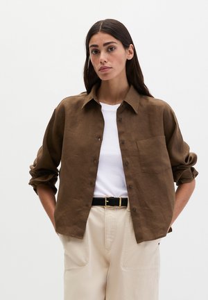 Žena s dlhými tmavými vlasmi, ktorá nosí hnedú košeľu na gombíky cez biele tričko, béžové nohavice a čierny opasok, stojaca na jednofarebnom pozadí.