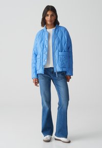 Veste matelassée, bleu clair, avec un col rond, des boutons-pression et deux poches avant, portée avec un jean bleu et des baskets blanches.