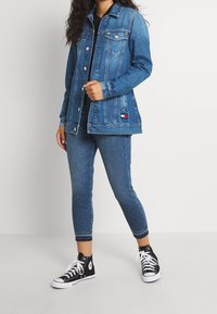 Chaqueta de mezclilla en azul medio, con un cuello clásico, bolsillos frontales y cierre de botones. Combinada con jeans azul oscuro y zapatillas negras de caña alta.