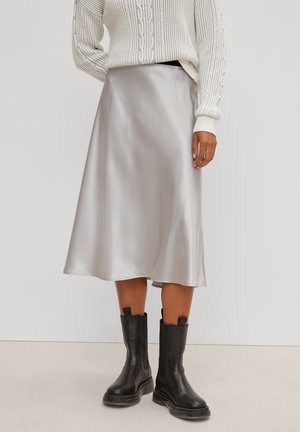 Femme portant un pull en maille torsadée blanc, une jupe midi en satin argenté et des bottes en cuir noires à mi-mollet sur un fond uni.