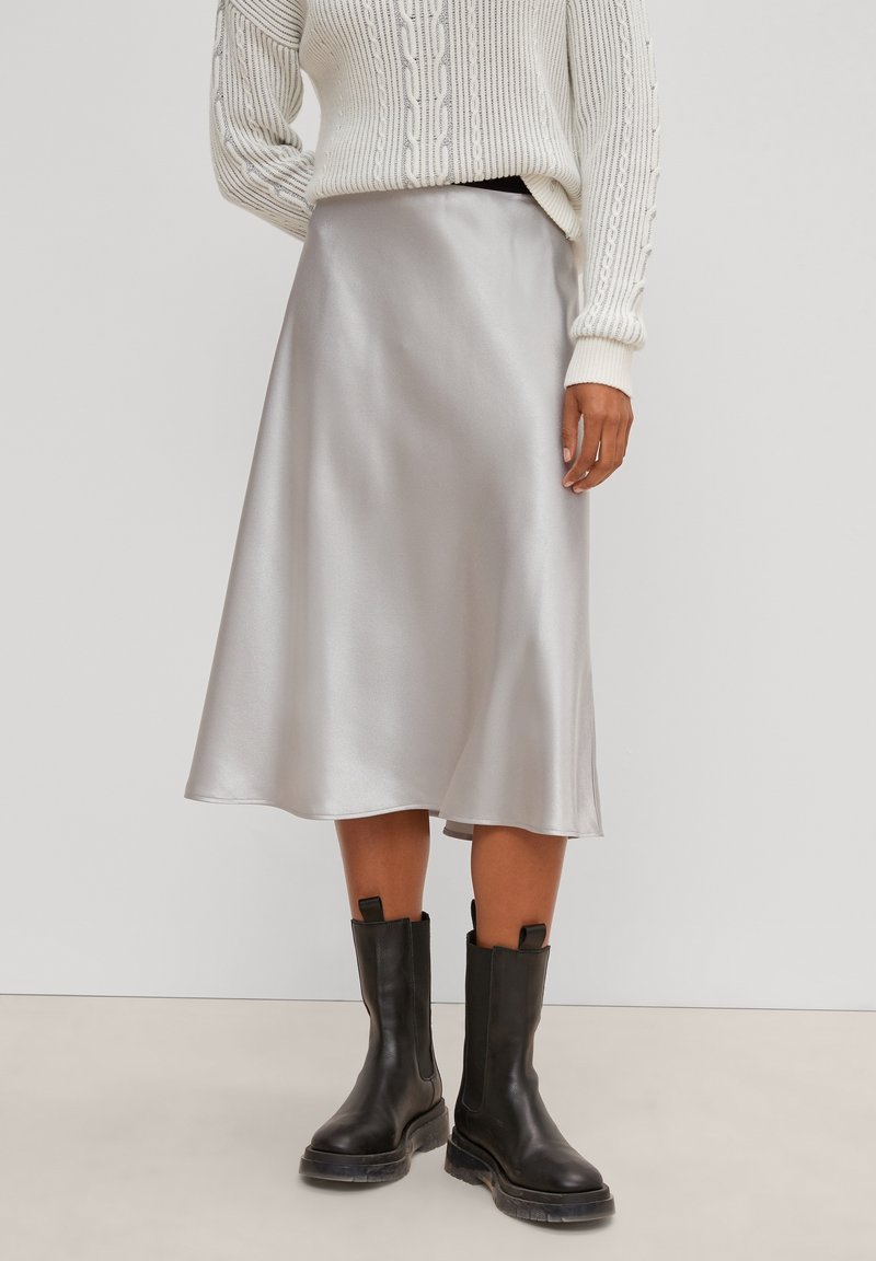Femme portant un pull en maille torsadée blanc, une jupe midi en satin argenté et des bottes en cuir noires à mi-mollet sur un fond uni.