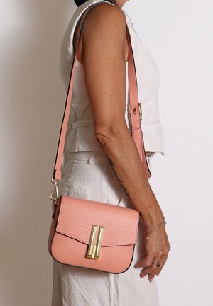 Donna con indosso abiti bianchi che porta una piccola borsa a tracolla rosa in pelle con chiusura dorata e tracolla regolabile.