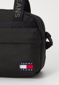 Sac en tissu noir avec fermeture éclair, doté d'un patch rectangulaire avec le logo "TOMMY JEANS" en blanc, rouge et bleu. Sangle réglable incluse.