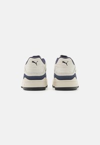 Le sneaker presentano un design bicolore con colori crema e blu scuro, una tomaia testurizzata, un collare imbottito e una suola in gomma nera. È visibile un logo.