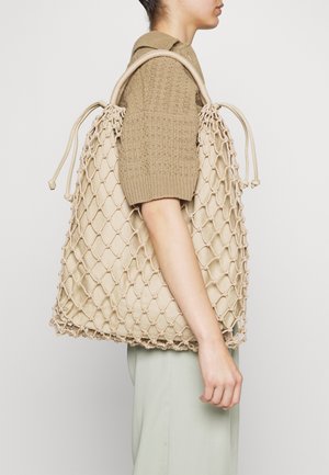 Osoba ubrana w beżowy, krótki sweter z dzianiny i jasne spodnie, niosąca duża beżową siatkowaną torbę na ramię z zamknięciem na sznurek.