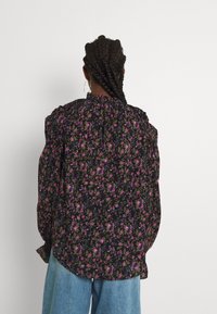 Svart blommig blus med lila och blå blommor, rynkade axlar och knäppning bak, tillsammans med ljusblå vida jeans.