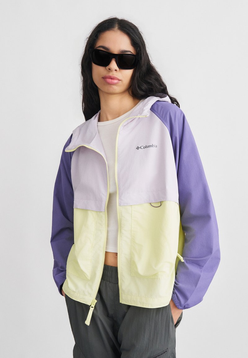 Γυναίκα με μακριά σκουρόχρωμα μαλλιά, φορώντας μαύρα γυαλιά ηλίου, ελαφρύ μωβ και κίτρινο μπουφάν Columbia, άσπρο κολλητό τοπ και σκούρο παντελόνι, σε απλό φόντο.