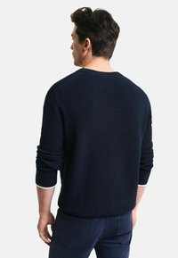 Granatowy sweter prążkowany o zorganizowanej konstrukcji, okrągłym dekolcie i kontrastujących białych akcentach przy mankietach, noszony przez mężczyznę z tyłu.