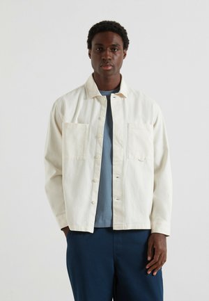 Uomo che indossa una camicia overshirt button-down off-white con due tasche sul petto, una maglietta blu sotto e pantaloni blu scuro su uno sfondo semplice.