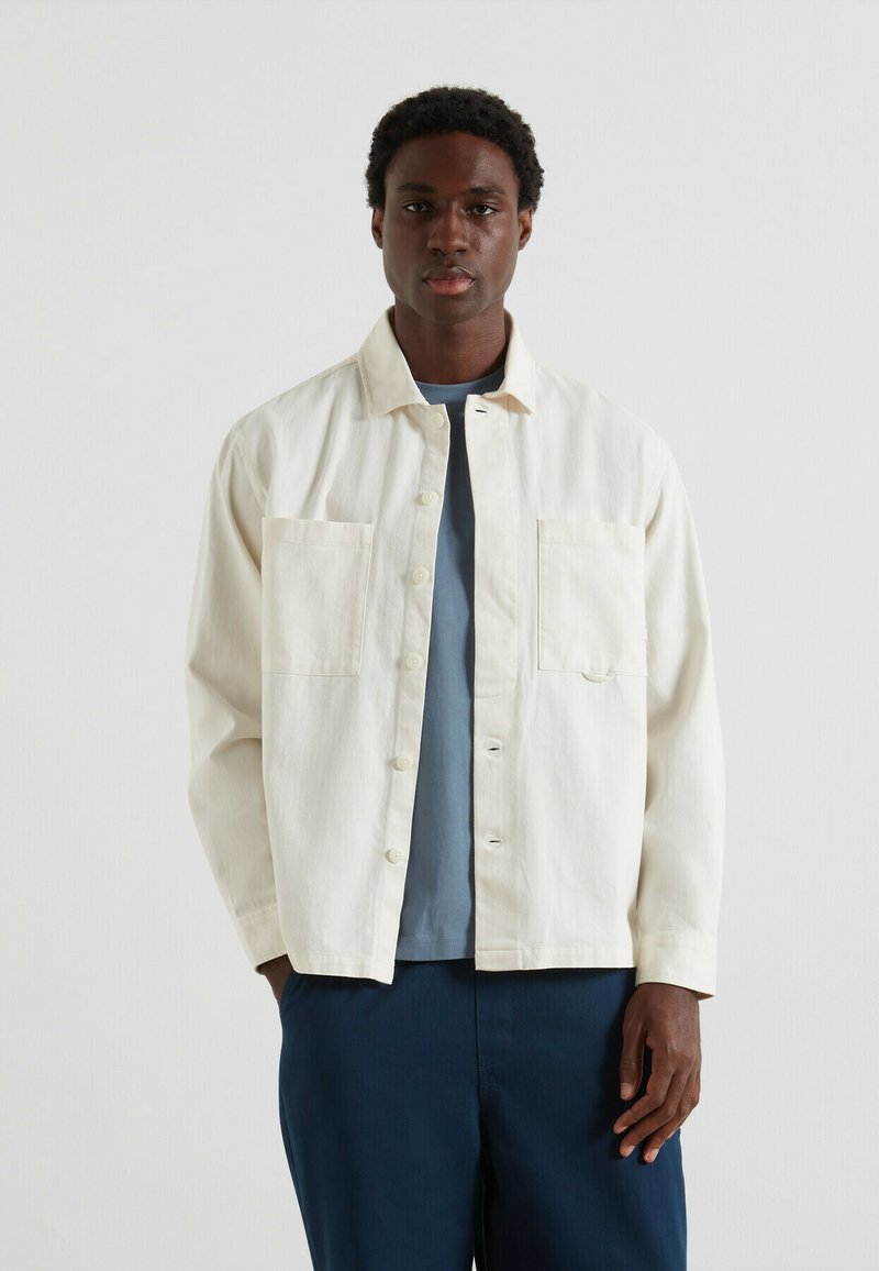 Uomo che indossa una camicia overshirt button-down off-white con due tasche sul petto, una maglietta blu sotto e pantaloni blu scuro su uno sfondo semplice.
