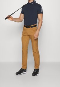 Man med marinblå polotröja och senapsgula byxor som håller en golfklubba vilande på axeln, står framför en enfärgad bakgrund.