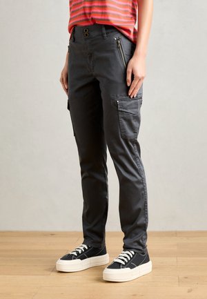 Person trägt dunkelgraue Cargo-Hose mit Reißverschluss- und Klappentaschen, schwarz-weiße Turnschuhe und ein rot gestreiftes Shirt, steht auf einem Holzfußboden.