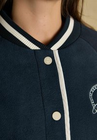 Tumes tekstuuriga jakk ribikrae, valgete aktsentide, tõmblukkude ja peene broderi kujundusega ees.