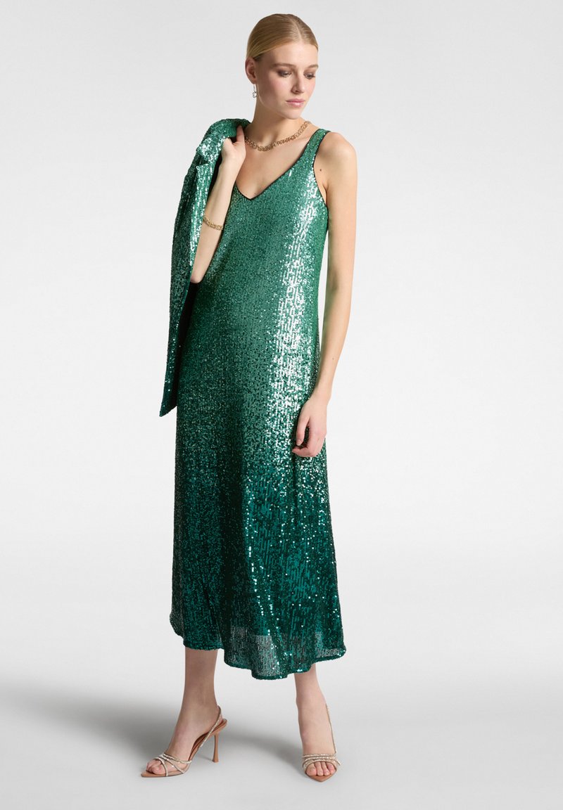 Abito verde con paillettes, scollo a V, lunghezza midi e silhouette aderente. Abbinato a una giacca coordinata. Tacchi a stiletto completano il look.