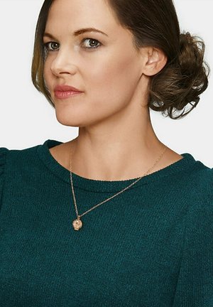 Collana in oro rosa con un pendente circolare, indossata su un maglione teal. La collana presenta una catena sottile che mette in risalto il design del pendente.