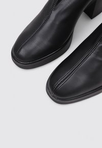Bottines en cuir noir avec des bouts carrés, des coutures verticales centrales et des talons bas sur un fond clair.