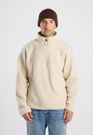 Crème-kleurige fleece pullover met een half-zip kraag, zijzak en een zachte textuur, gecombineerd met een bruine geribde gebreide beanie.