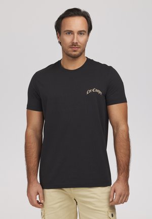 Mężczyzna w czarnej koszulce Lee Cooper i beżowych spodniach typu cargo, stojący na jednolitym jasnoszarym tle, zwrócony twarzą do przodu.