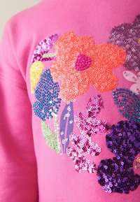 Pull rose vif avec des fleurs pailletées multicolores en orange, violet, bleu, jaune et vert sur la poitrine.