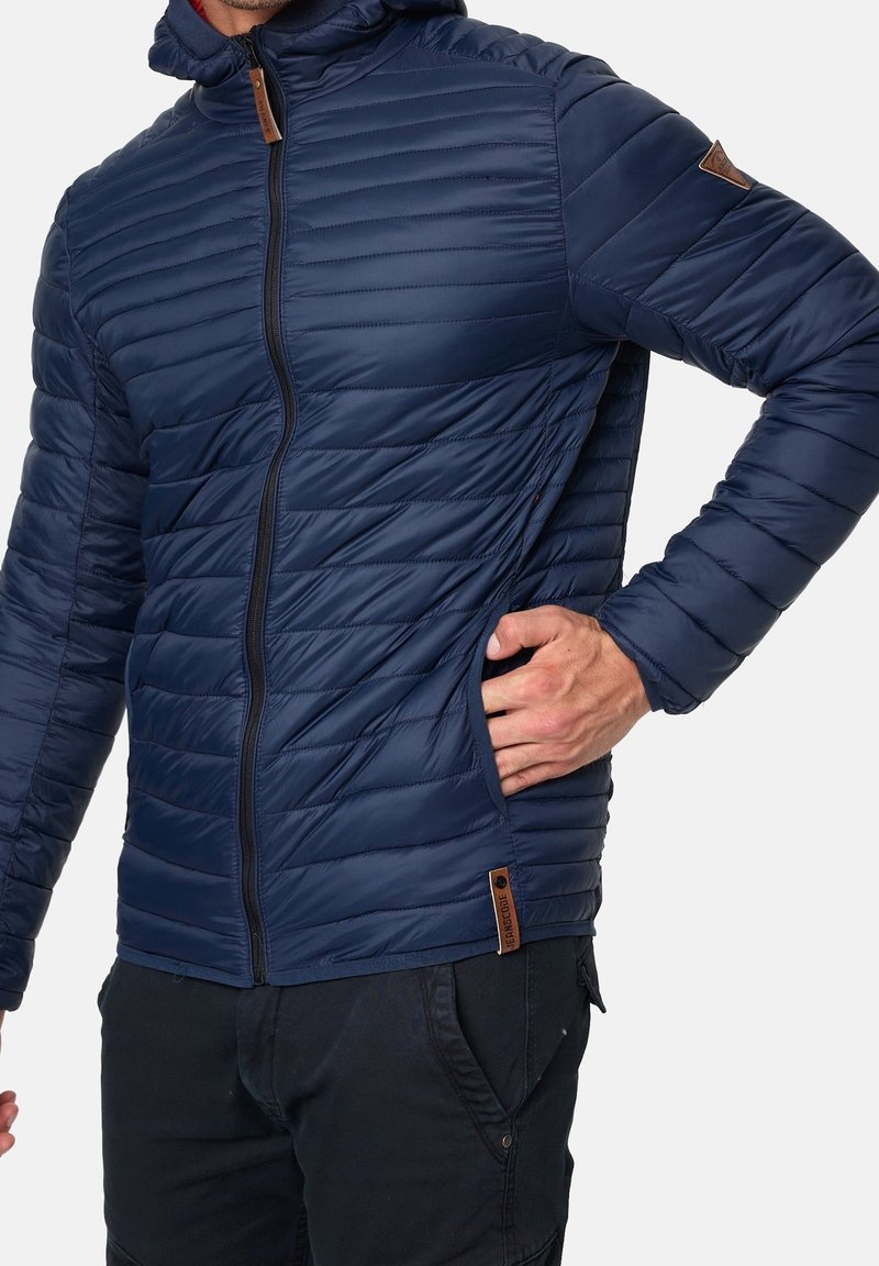 Navy blue padded jacket featuring horizontal quilted design, full zip front, hood, and leather accent on sleeve; side pocket visible.