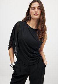 Sort draperet top lavet af letvægtsstof med en skulderdesign og sammensat tekstur, parret med løse bukser. Sølvarmbånd som accent.