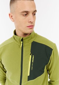 Jonge man in een groene fleecejack met een zwarte ritssluiting, gelabeld "Protest Pyre Green," kijkend naar links.