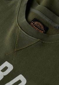 Sudadera verde oliva con un escote redondo, que presenta un destacado estampado blanco y un detalle de costura en cruz cerca del cuello. Etiqueta de la marca visible.