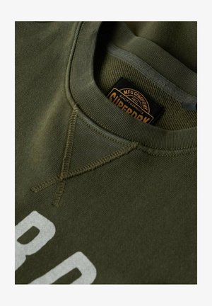 Sudadera verde oliva con un escote redondo, que presenta un destacado estampado blanco y un detalle de costura en cruz cerca del cuello. Etiqueta de la marca visible.