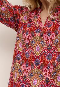 Kvinna i en röd blus med paisleymönster och detaljer i rosa, blått och guld, visar upp halsringningen och höjd arm.