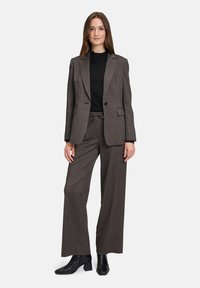 Braun karierter Blazer über einem schwarzen Oberteil, kombiniert mit locker sitzenden braun karierten Hosen. Das Model trägt schwarze Knöchelstiefel.