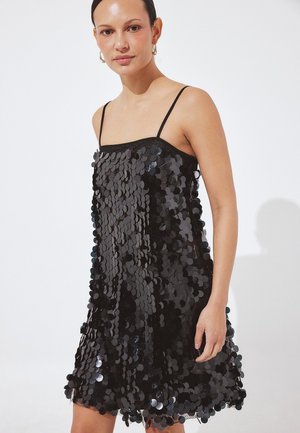 Vrouw die een mouwloze zwarte jurk draagt bedekt met grote glanzende pailletten, staand tegen een effen witte achtergrond met gouden oorringen.