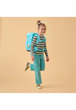 Junges Mädchen trägt türkise Cordhosen, gestreiften Pullover, beigen Sneaker und einen großen blauen Rucksack, steht mit einem angehobenen Bein vor einem beigen Hintergrund.