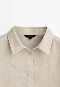 Chemise en lin beige avec col pointu et deux boutons visibles sur la patte de boutonnage avant, montrée de la partie supérieure de la poitrine jusqu'au col.