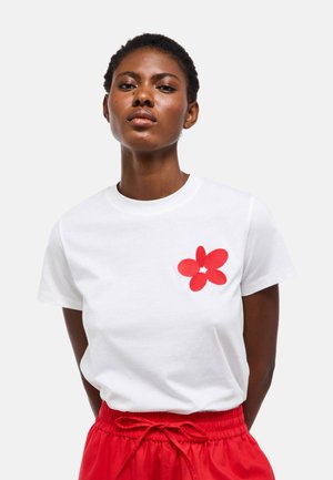 Femme aux cheveux courts et bouclés portant un T-shirt blanc avec un motif de fleur rouge et un pantalon rouge à cordon, debout devant un fond uni.