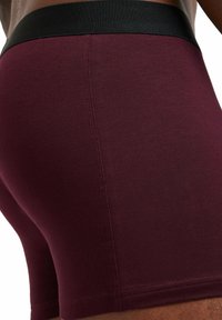 Boxer shorts bordeaux avec une taille élastique noire, fabriqués à partir d'un tissu lisse, présentant une couture soignée et un design ajusté.