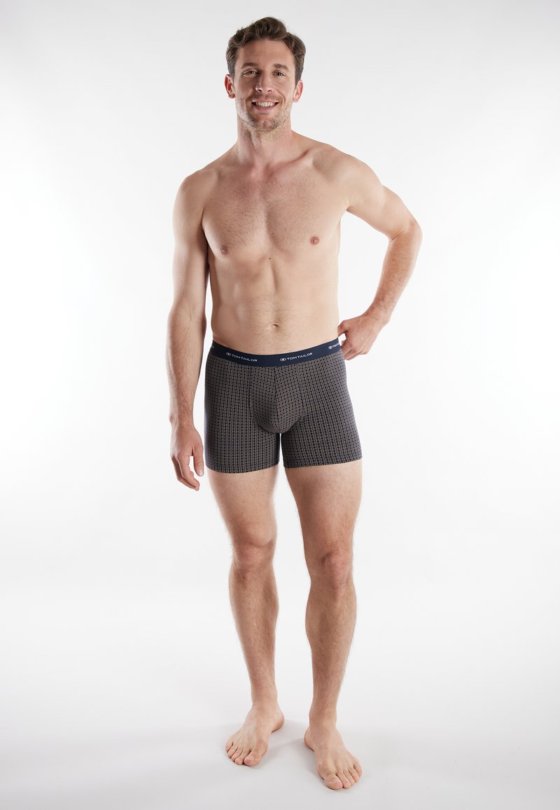 Men's fitted boxer shorts in navy with a geometric pattern, soft fabric, and elastic waistband featuring a brand logo. Model standing barefoot.