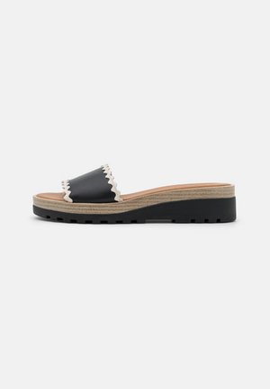 Black slide sandal with a scalloped edge strap, natural-colored midsole, and textured black rubber outsole. Casual, platform design.