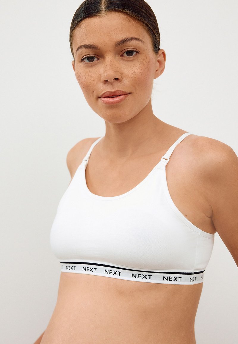 Vrouw met sproeten die een witte sportbeha draagt met verstelbare bandjes en een merkgebande elastische band met de tekst "NEXT" rond de borst.