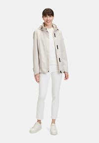 Veste imperméable beige avec capuche, zips et accents noirs, portée sur un haut blanc, assortie à un pantalon blanc et des baskets légères.