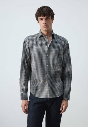 Joven de pie vistiendo una camisa de manga larga azul y beige a cuadros con botones y pantalones oscuros sobre un fondo claro liso.