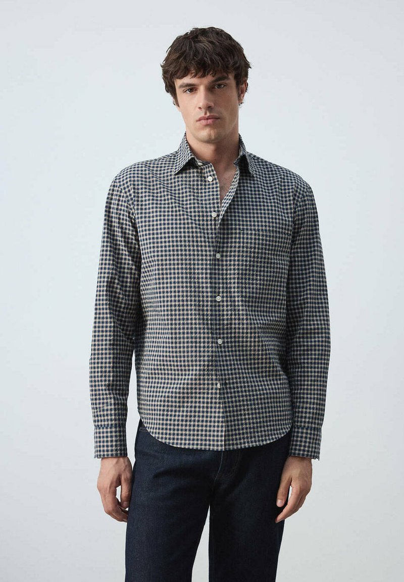 Jeune homme debout portant une chemise à manches longues à carreaux bleu et beige boutonnée et un pantalon foncé sur un fond clair uni.