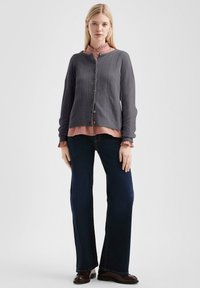 Grijze gebreide cardigan met verticale lijnen, bruine knopen, gelaagd over een roze blouse met ruches, gecombineerd met donkerblauwe flared jeans.