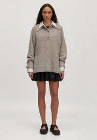 Hellbrauner Strickpullover mit einem darunter getragenen Kragenhemd, kombiniert mit einem schwarzen, Rüschensrock und dunklen Sneakers. Weicher Stoff und lässige Passform.