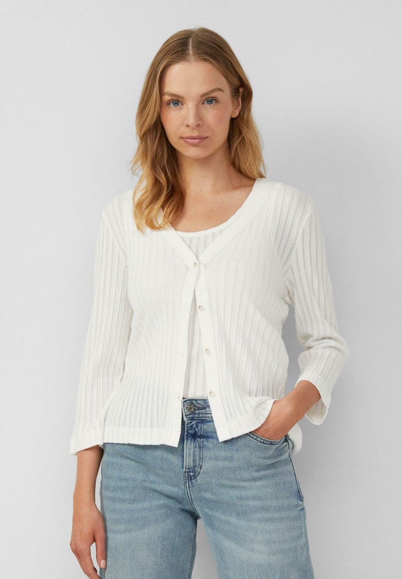 Witte geribbelde cardigan met een V-hals, met knoopsluiting en driekwartmouwen, gedragen over een lichte top en blauwe jeans.
