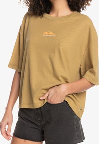 Kortärmad, senapsgul t-shirt i våffeltextur med logodetalj på bröstet, parat med svarta shorts, avslappnad passform och fåll.